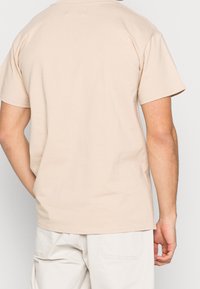 Muž v béžovém tričku s krátkým rukávem a světlých kalhotách, zobrazený zezadu s jednou rukou spočívající vedle těla.