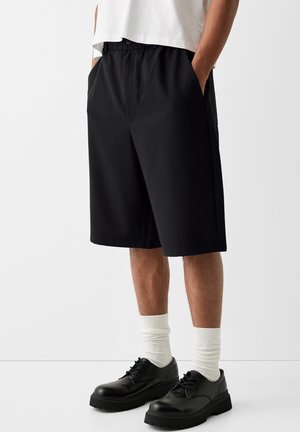 Homme portant un short noir jusqu'aux genoux, des chaussettes blanches à hauteur de mollet, des chaussures noires à lacets et un t-shirt blanc court, debout les mains dans les poches.