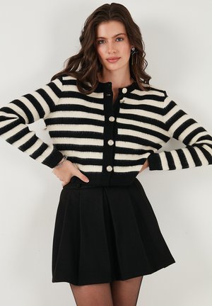 Cardigan a righe bianche e nere, lavorato a maglia, con scollatura rotonda e chiusura a sei bottoni. Indossato sopra una gonna nera plissettata. Texture liscia.