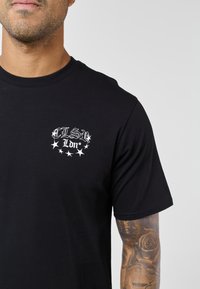 Μαύρο βαμβακερό t-shirt με λευκό γραφικό τύπωμα στο στήθος, με γράμματα και λεπτομέρειες με αστέρια. Κοντά μανίκια και λαιμόκοψη στρογγυλή.