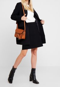 Cappotto nero sopra una maglietta bianca, abbinato a una gonna nera. Borsa a tracolla in pelle marrone con dettaglio a frange e stivaletti neri con tacco.