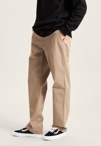 Pantaloni in cotone beige con tasche laterali, abbinati a sneakers bianco e nere con tomaia in suede e suola in gomma bianca.