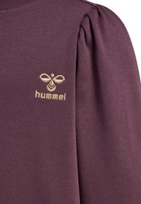 Fioletowa bluza z puff sleeves, ryżowym kołnierzykiem oraz złotym haftowanym logo z motywem pszczoły na lewej piersi. Gładka faktura materiału.