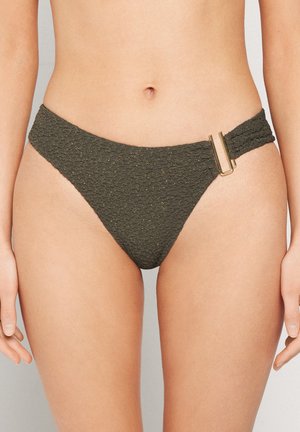 Kobieta nosząca teksturowane ciemnozielone spodnie bikini z prostokątną złotą sprzączką na prawym biodrze, na neutralnym tle.