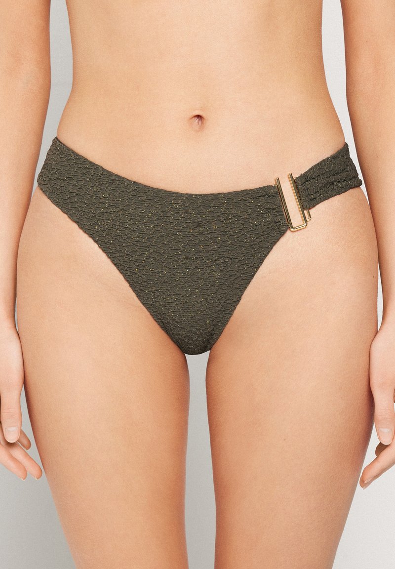 Donna che indossa slip da bikini verde scuro testurizzato con fibbia rettangolare dorata sull'anca destra, su sfondo neutro.