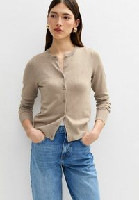 Beige cardigan med rund halsringning, framknäppning och ribbade ärmslut. Långt tillsammans med blå jeans, med händerna i fickorna, på en ljus bakgrund.
