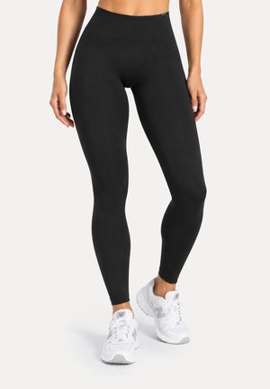 Femme portant un legging noir taille haute et des baskets blanches New Balance, debout devant un fond clair uni.