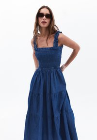 Sininen farkku maksimekko, jossa on kiristettävä yläosa, röyhelöolkaimet ja kerrostettu hameosa. Liukas materiaali ja rento istuvuus, suunniteltu mukavuutta varten.