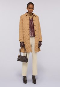 Cappotto trapuntato marrone con bottoni neri, top in pelle bordeaux, pantaloni crema, guanti marroni e una piccola borsa scura. Stivaletti con tacco.