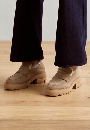 Brune ruskinds loafers med en tyk, tekstureret gummisål, der har en blød indvendig foring og et enkelt bånd over forsiden.