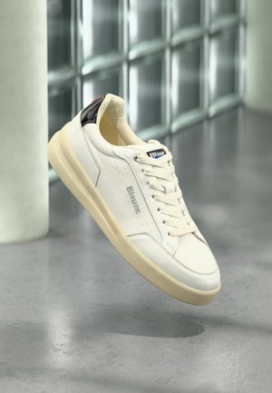 Witte leren sneaker met een crème rubberen zool, zwarte hielaccent en beige veters. Voorzien van logo en minimale perforaties aan de zijkant.