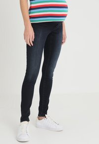 Materniteit skinny jeans in donkerblauwe denim. Heeft een rekbare buikband, aansluitende benen en een schone afwerking. Gecombineerd met witte sneakers.