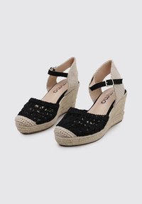 Semelles compensées en espadrilles tissées noires avec des brides de cheville tissées beiges, présentent un design texturé et des détails en corde sur une semelle en jute.