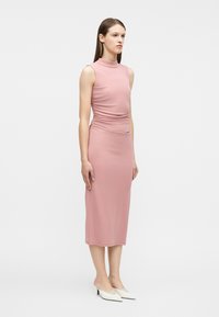 Γυναίκα φορώντας ροζ μίντι φόρεμα χωρίς μανίκια και με ψηλό λαιμό, με ραφές στη μέση, και λευκά μίνι μυτερά τακούνια, στέκεται μπροστά σε απλό φόντο.