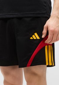 Črne atletske kratke hlače z rumenim logotipom Adidas, rdečimi in rumenimi črtami na strani ter gladko teksturo. Roka je položena na pasu.