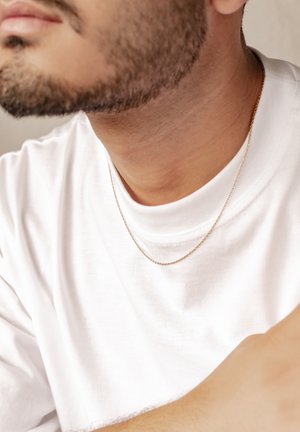 Gold chain necklace resting on a bare skin neck. The wearer is in a white t-shirt, with visible facial hair and slight shadowing.