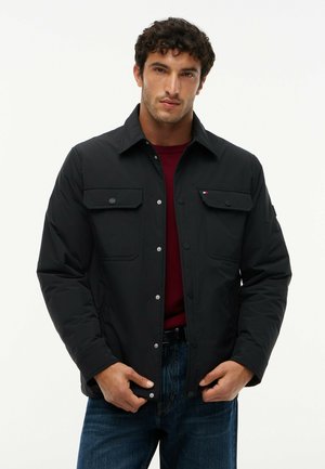 Mand iført sort knapjakke over en vinrød skjorte og blå jeans, står og knapper jakken til mod ensfarvet baggrund.