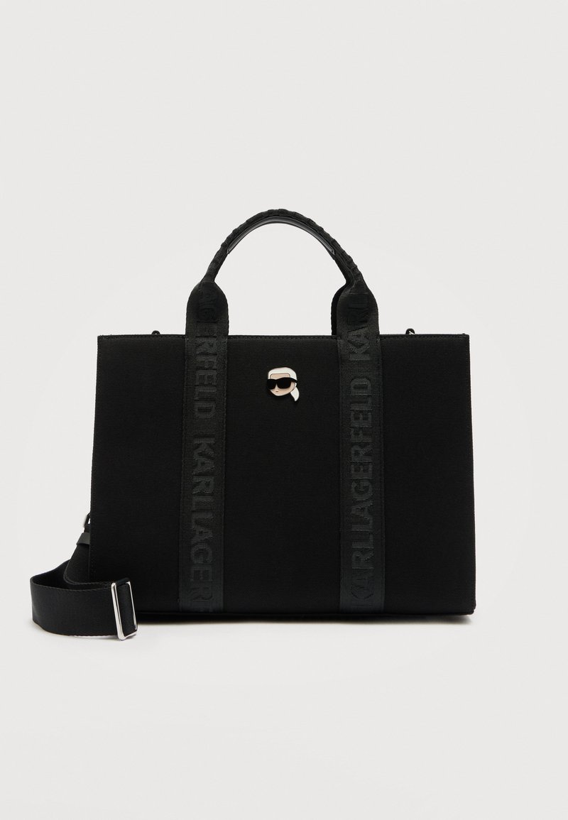 Μαύρη ορθογώνια τσάντα tote με δύο χερούλια, ρυθμιζόμενο λουρί ώμου και μια μικρή καρφίτσα με τη φιγούρα ενός ατόμου που φορά γυαλιά ηλίου στο μπροστινό μέρος.