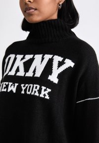 Svart stickad tröja med hög ribbad krage, med en iögonfallande textdesign i vitt som säger "DKNY NEW YORK". Strukturerad tyg med subtila vita sömmar.