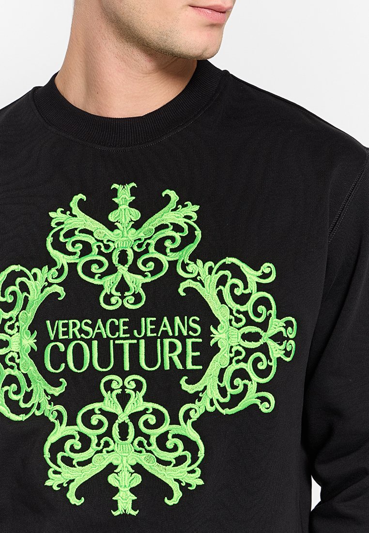 Sudadera negra con intrincados bordados en verde de un diseño decorativo y el texto "VERSACE JEANS COUTURE" en negrita en el centro.