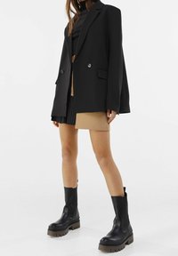 Överdimensionerad svart blazer med två knappar, lagd över en brun minikjol. Svarta ankelstövlar med tjocka sulor fulländar outfiten.