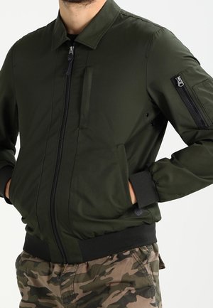 Homme portant une veste bomber vert foncé avec des poches zippées et un pantalon cargo camouflage, les mains dans les poches de la veste.