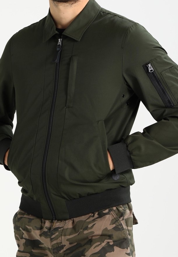 Homme portant une veste bomber vert foncé avec des poches zippées et un pantalon cargo camouflage, les mains dans les poches de la veste.