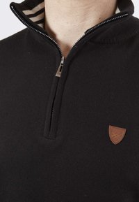 Pull noir en tricot avec un col zippé, doté d'un col interne rayé et d'une petite patch en cuir marron avec un symbole à l'avant.