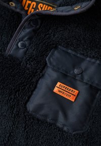 Fleece pullover i mørk marineblå med en struktureret, plysset finish. Har en brystlomme med et orange "Superdry MFG/CO" mærke og tryklukninger.