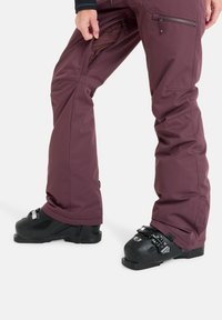 Bordeauxkleurige skibroek met een relaxte pasvorm, voorzien van ritssluitingen en een gestructureerde afwerking, gecombineerd met zwarte skischoenen.