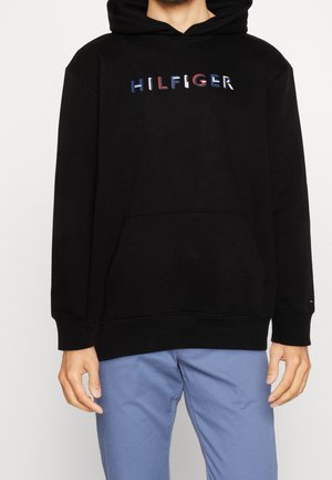 Sort hættetrøje med flerfarvet "HILFIGER" tekst på brystet, båret af en person med lyseblå bukser, hænder afslappet ved siderne.