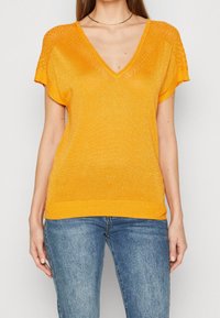 T-shirt z dekoltem w serek w jaskrawym pomarańczowym kolorze, z krótkimi rękawami i teksturowanym koronkowym wykończeniem na ramionach. Miękka, połyskująca tkanina z prążkowanym dołem.