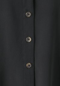 Camisa de tela negra con una textura suave, que presenta cinco botones y costuras limpias a lo largo de la tapeta. Los botones son redondos con un sutil brillo.