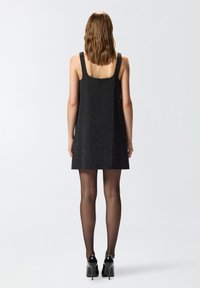 Vestido negro sin mangas con un escote cuadrado, que presenta un patrón texturizado. Combinado con medias negras transparentes y zapatos de tacón alto brillantes.