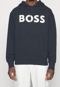 Muž v tmavě modré mikině s kapucí a bílým nápisem "BOSS" přes hrudník a v bílých kalhotách, stojící na jednobarevném pozadí.