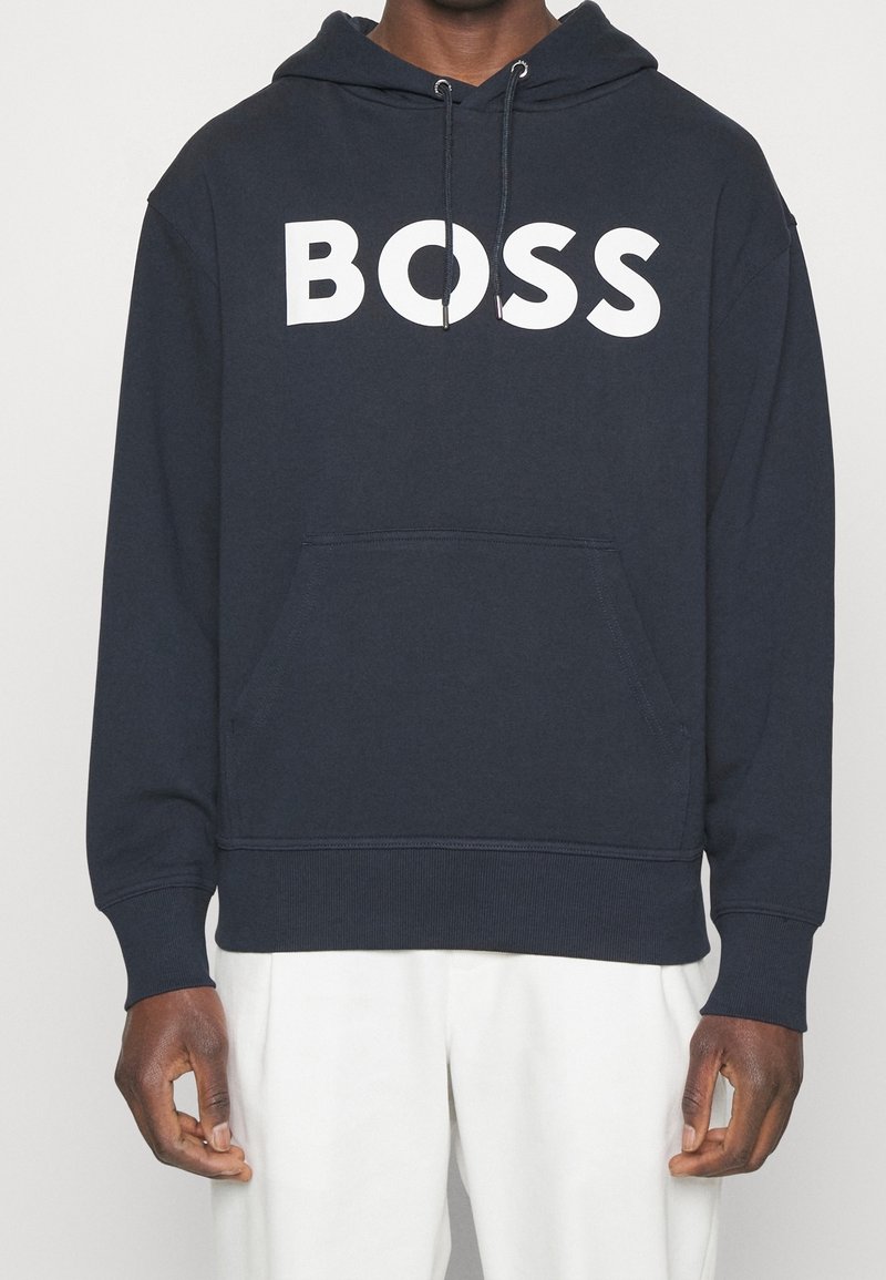 Muž v tmavě modré mikině s kapucí a bílým nápisem "BOSS" přes hrudník a v bílých kalhotách, stojící na jednobarevném pozadí.