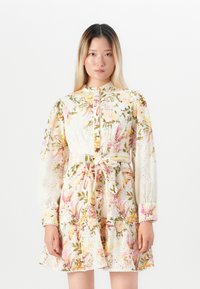 Donna che indossa un abito lungo con maniche, con cintura, a motivo floreale nei toni del rosa, giallo e verde su sfondo bianco, che si trova davanti a uno sfondo bianco uniforme.