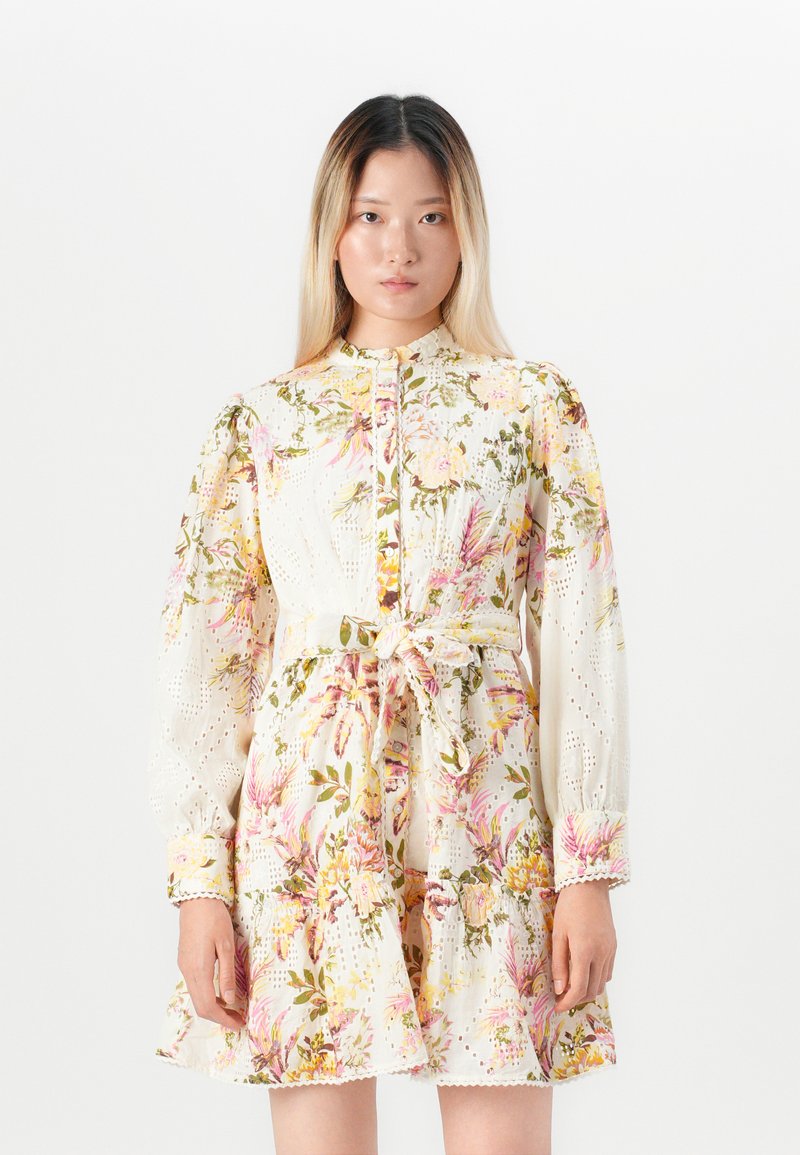 Donna che indossa un abito lungo con maniche, con cintura, a motivo floreale nei toni del rosa, giallo e verde su sfondo bianco, che si trova davanti a uno sfondo bianco uniforme.