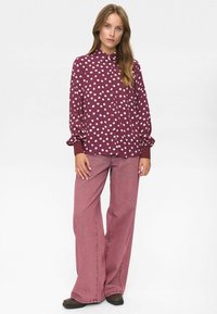 Femme portant une chemise boutonnée à manches longues bordeaux avec des pois blancs et un pantalon en jean rose à jambes larges, se tenant devant un fond uni.