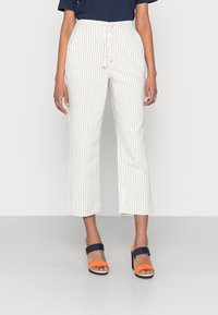 Pantalon blanc à rayures avec une taille nouée, longueur mi-mollet, associé à des sandales à talons blocs orange et marine, texture de tissu lisse.