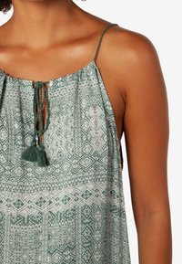 Buste d'une personne à la peau brun moyen portant une robe sans manches, verte et blanche à motifs, avec des bretelles fines et un lien à pompons au niveau du col.