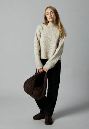 Pull en laine crème avec un motif en relief, un pantalon loose-fit noir et un sac en suédine marron porté à la main. Le pull a un col rond et des poignets côtelés.