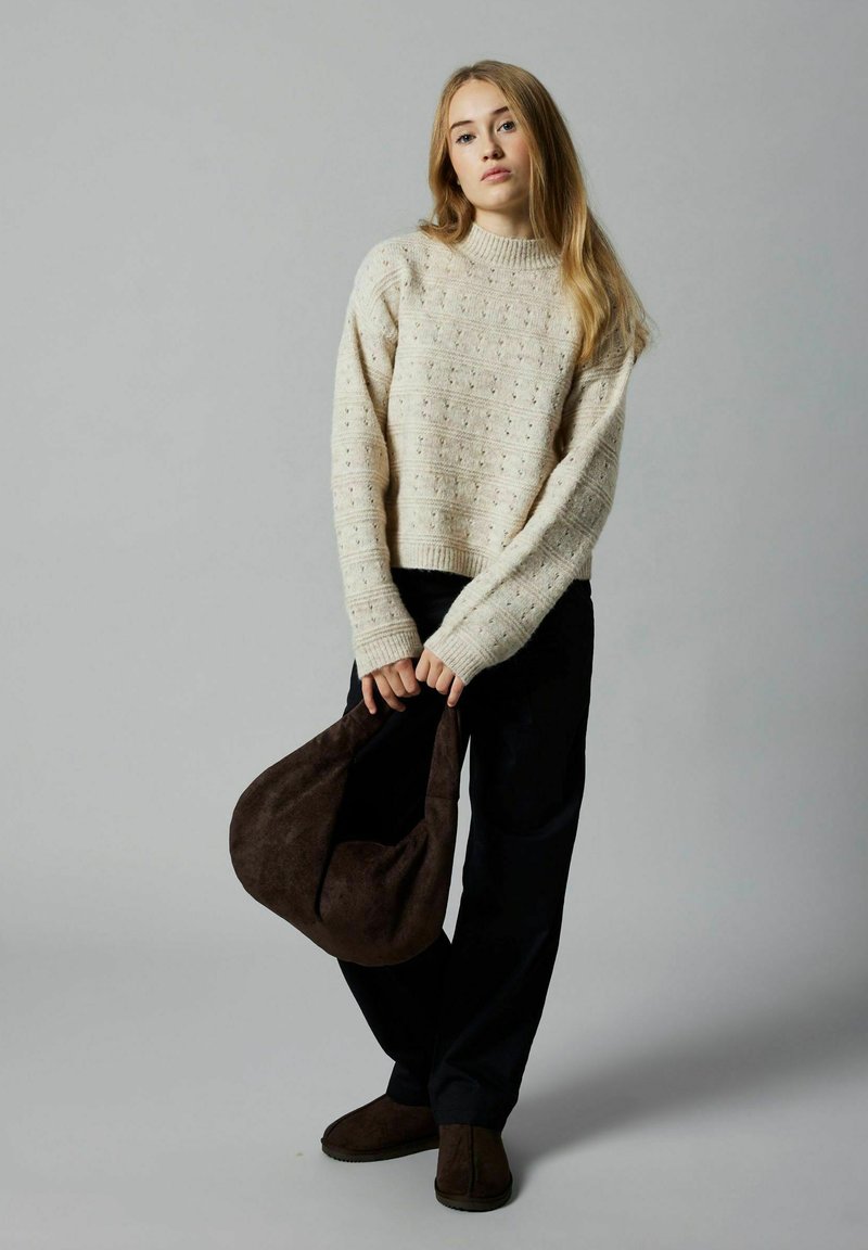 Cremfarbener, gestrickter Pullover mit erhobenem Muster, dunkel lockere Hose und eine braune Wildledertasche in der Hand gehalten. Der Pullover hat einen runden Ausschnitt und gerippte Bündchen.
