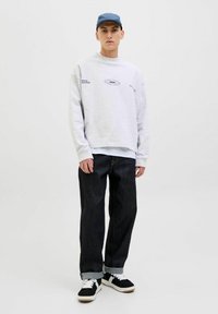 Sudadera gris con logo, superpuesta a una camisa blanca. Vaqueros oscuros con dobladillo. Zapatillas negras con detalles y cordones blancos. Gorra azul.