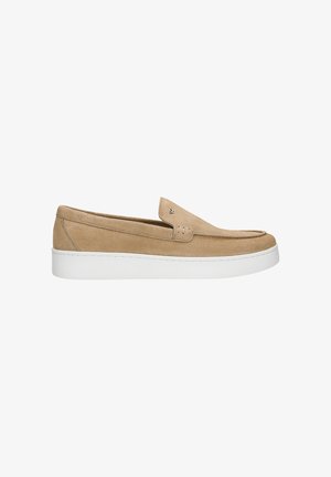 Man staat met handen in de zakken, draagt een beige polo met korte mouwen, lichtblauwe spijkerbroek, beige slip-on schoenen en zichtbare tatoeages op de armen.