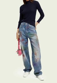 Jasnoniebieskie jeansy dżinsowe z wielokolorowymi plamami farby, luźny krój, lekko postrzępione na kolanach, zestawione z ciemnym dzianinowym swetrem i białymi sneakersami.