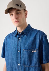 Sötétkék farmering rövid ujjakkal, mellzsebbel és Calvin Klein logóval. Kiegészítők között szerepel egy bézs sapka.