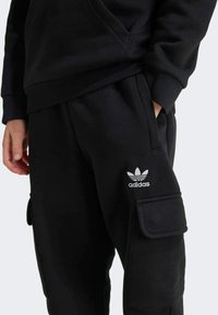 Čierna mikina so predným vreckom a tepláky s bočnými cargo vreckami. Logo Adidas v bielej farbe na teplákach. Materiál z bavlnenej zmesi, voľný strih.