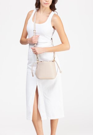 Donna con canotta bianca e gonna che tiene una borsa a tracolla in pelle beige con tracolla regolabile e chiusura a coulisse.