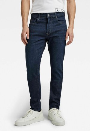 Homme portant un jean slim bleu foncé et des baskets blanches, visible de la taille aux pieds sur fond blanc.